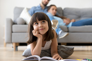 Young girl reading a book on the floor with a couple resting on the couch behind her